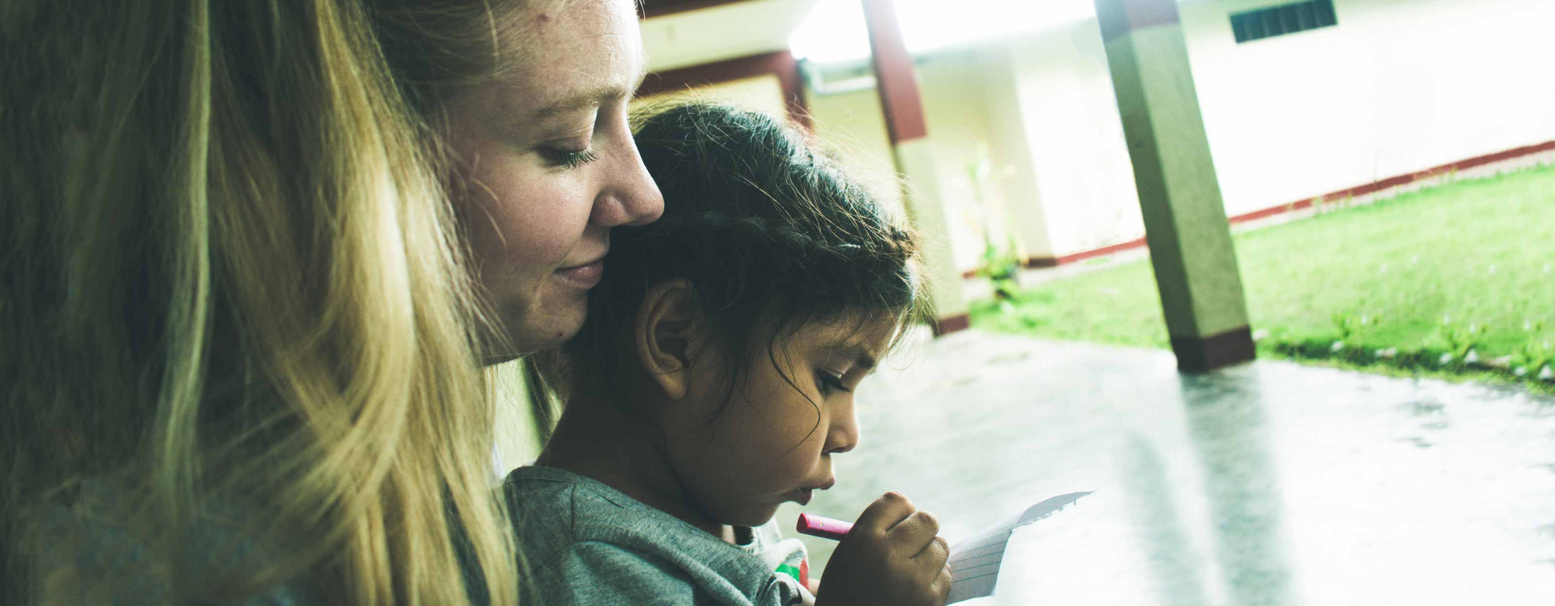 A student looks over a young girl's shoulder as she writes on a piece of paper