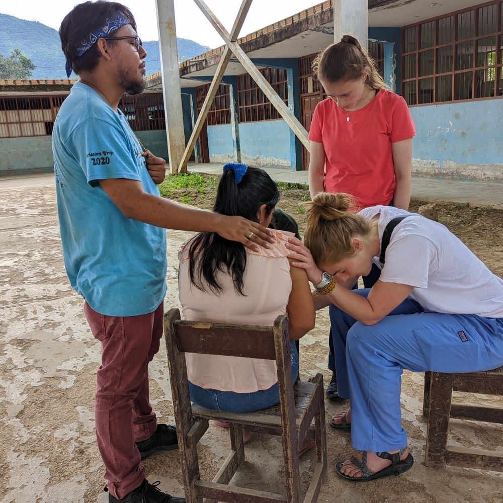 A group of people pray over a seated person, with two of them putting their hands on the person's shoulder