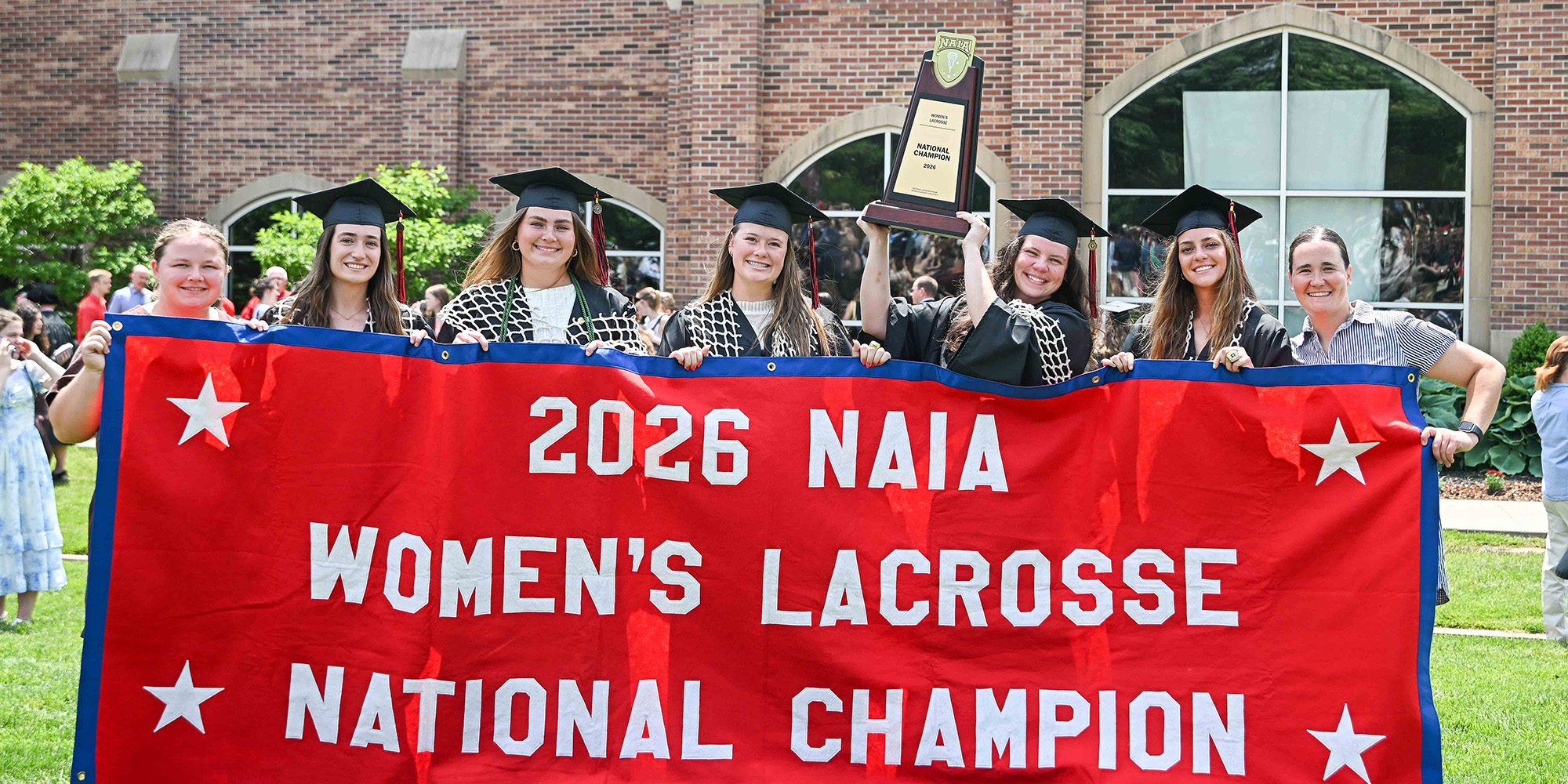 Graduates from the 2026 NAIA Women's Lacrosse National Championship winning team pose for a photo with their trophy and victory banner after Commencement
